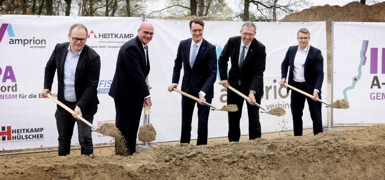 Das Foto zeigt fünf Männer in Anzügen, die an einem Spatenstich teilnehmen. Sie stehen nebeneinander an einer länglichen, aufgeworfenen Erdgrube und halten goldfarbene Spaten in den Händen, mit denen sie Erde bewegen. Im Hintergrund ist ein weißes Banner mit verschiedenen Logos und Schriftzügen zu sehen. Links ist das Amprion-Logo mit dem Schriftzug "amprion verbindet" erkennbar. Daneben steht "HEITKAMP" mit dem Untertitel "Baumannschaften. Unsere Welt. Unsere Leistung.". Rechts sind Teile eines weiteren Logos mit "A-" und "GEM" sichtbar. Hinter dem Banner befinden sich Bäume ohne Blätter und ein sandiger oder erdfarbener Hügel. 