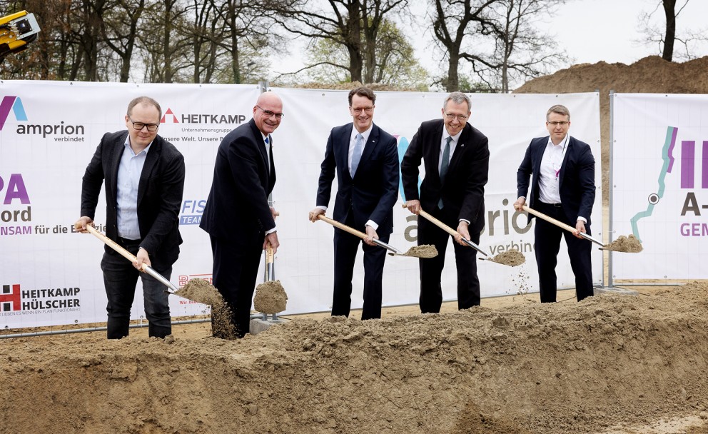 Das Foto zeigt fünf Männer in Anzügen, die an einem Spatenstich teilnehmen. Sie stehen nebeneinander an einer länglichen, aufgeworfenen Erdgrube und halten goldfarbene Spaten in den Händen, mit denen sie Erde bewegen. Im Hintergrund ist ein weißes Banner mit verschiedenen Logos und Schriftzügen zu sehen. Links ist das Amprion-Logo mit dem Schriftzug "amprion verbindet" erkennbar. Daneben steht "HEITKAMP" mit dem Untertitel "Baumannschaften. Unsere Welt. Unsere Leistung.". Rechts sind Teile eines weiteren Logos mit "A-" und "GEM" sichtbar. Hinter dem Banner befinden sich Bäume ohne Blätter und ein sandiger oder erdfarbener Hügel.