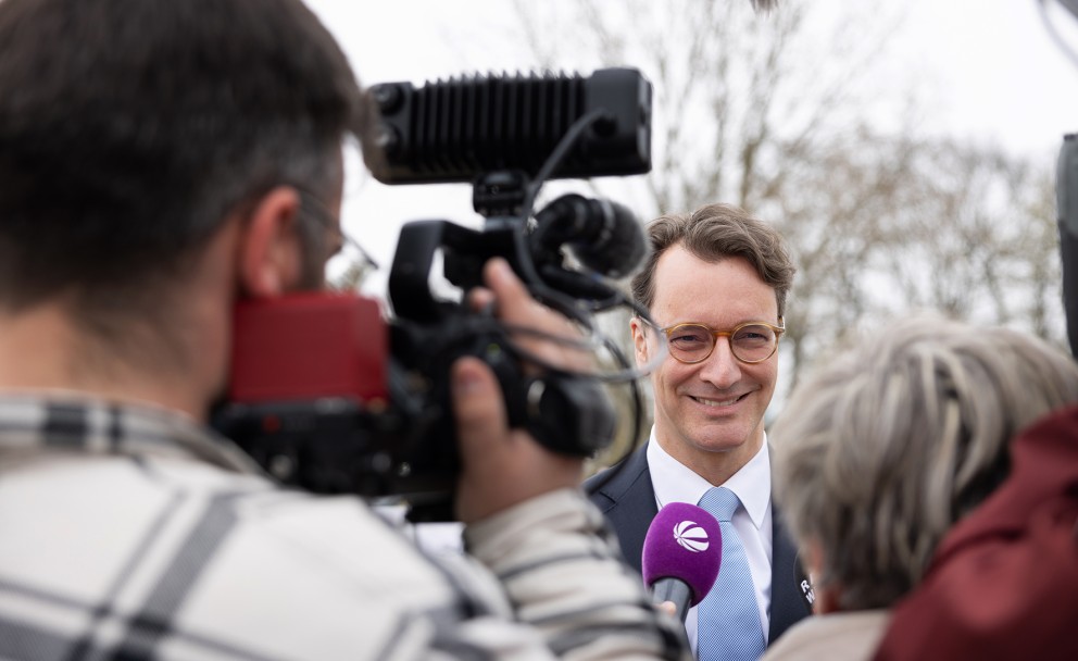 Das Bild zeigt eine Nahaufnahme eines Mannes mit hellbraunen, leicht gewellten Haaren und Brille, der in die Kamera lächelt. Er trägt einen dunklen Anzug, ein weißes Hemd und eine hellblaue Krawatte mit einem dezenten Muster. Vor seinem Mund ist ein lilafarbenes Mikrofon mit einem weißen Logo platziert, das wie ein stilisiertes "e" aussieht. Das Mikrofon wird von einer Person gehalten, die außerhalb des Bildrahmens zu sein scheint.
Im Vordergrund links ist der Rücken einer Person mit dunklen, kurzen Haaren zu sehen, die eine Videokamera bedient. Die Kamera ist schwarz mit roten Akzenten und einem großen Objektiv. Ein Mikrofon mit einem Windschutz ist oben an der Kamera befestigt und ragt in den oberen Bildrand.
Rechts im Vordergrund ist der Hinterkopf einer Person mit kurzen, grauen Haaren zu sehen, die dem Mann mit dem Mikrofon zuzuhören scheint.
Der Hintergrund ist unscharf und zeigt helle, möglicherweise bewölkte oder neblige Bedingungen mit verschwommenen Bäumen oder Strukturen.