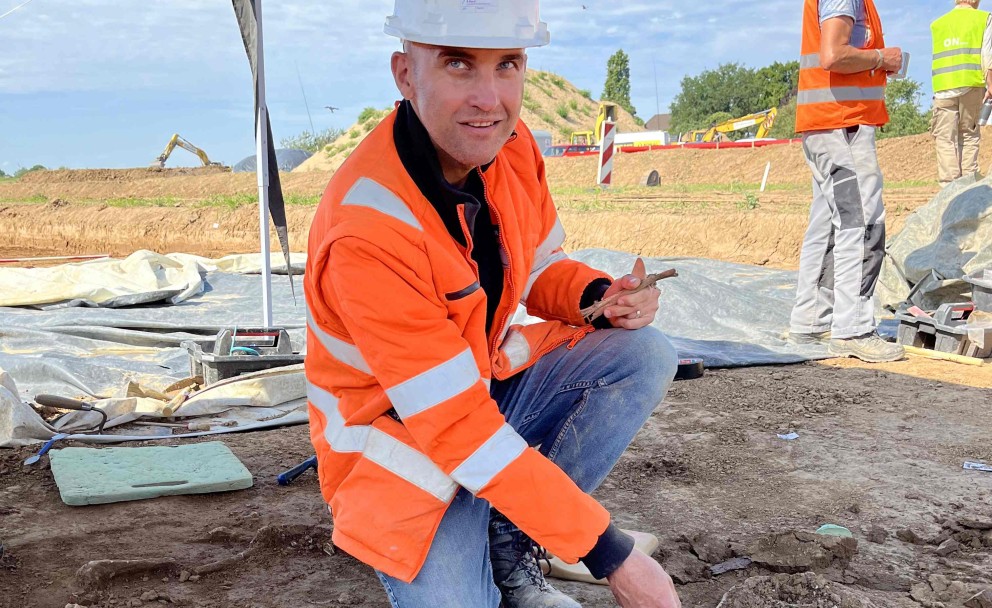 Auf dem Foto kniet ein Mann in orangefarbener Warnweste und einem weißen Schutzhelm auf einer archäologischen Ausgrabungsstätte. Er hält einen Stab in der Hand, mit dem er vorsichtig auf eine freigelegte Stelle im Boden zeigt. Direkt vor ihm sind die Knochen eines menschlichen Skeletts zu erkennen, das teilweise von Erde bedeckt ist. Im Hintergrund steht ein weiterer Arbeiter, ebenfalls mit Warnweste und Schutzhelm, und eine teilweise abgedeckte Fläche mit Planen ist sichtbar.